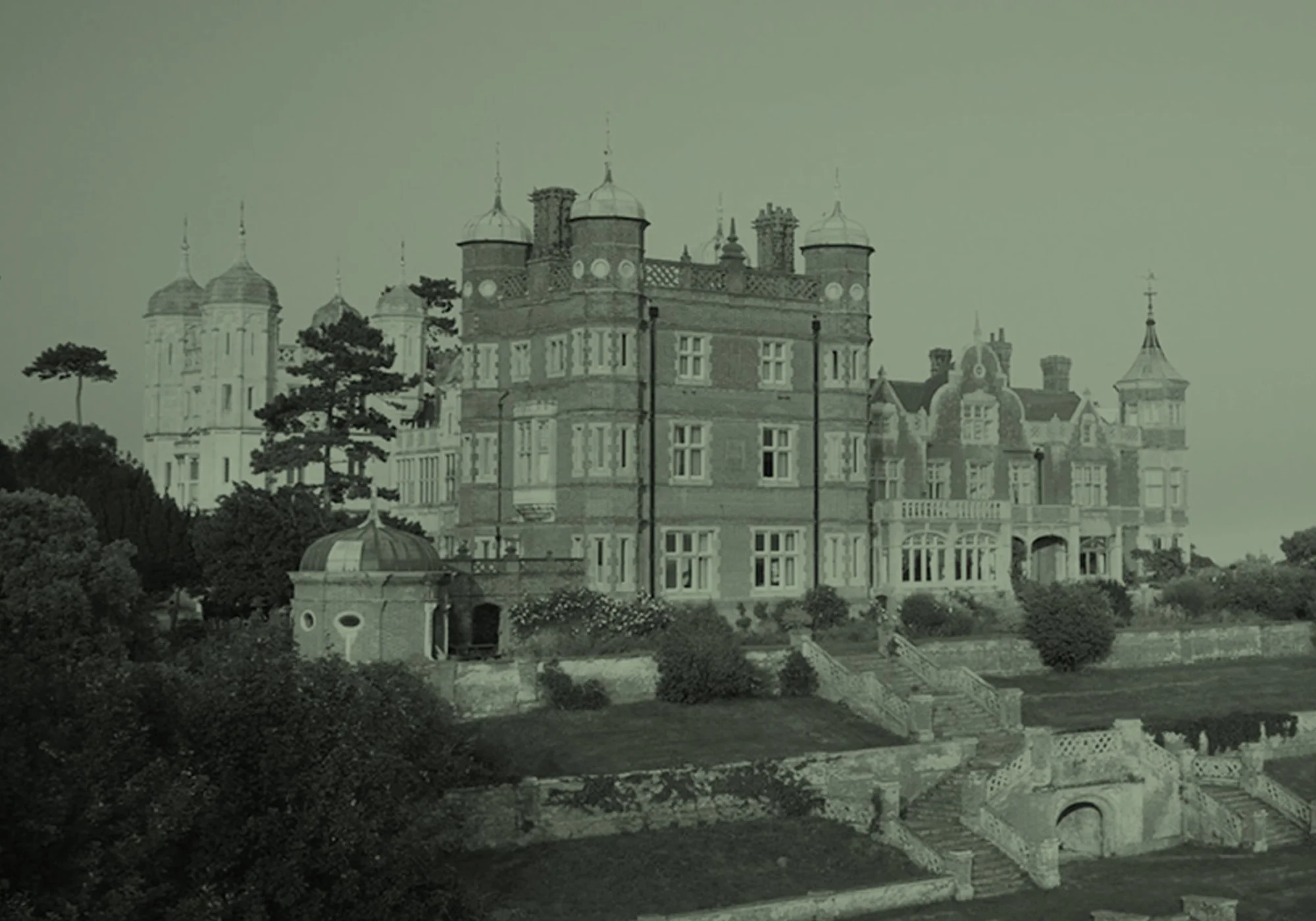 A black and white photo of a large, ornate castle with multiple turrets, surrounded by trees and a garden with pathways.