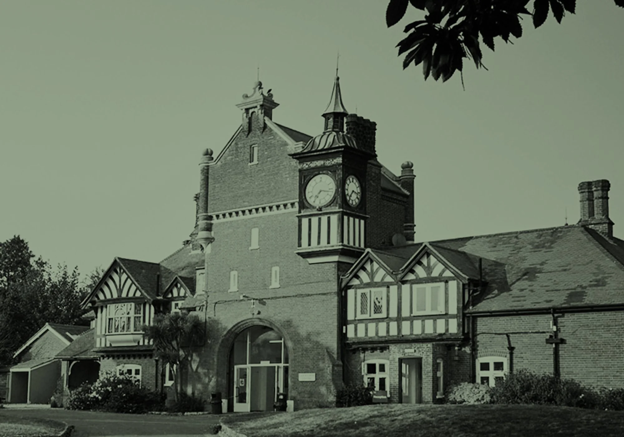 A historic brick building with a clock tower, steep roofs, and Victorian architectural details, surrounded by trees and a lawn.