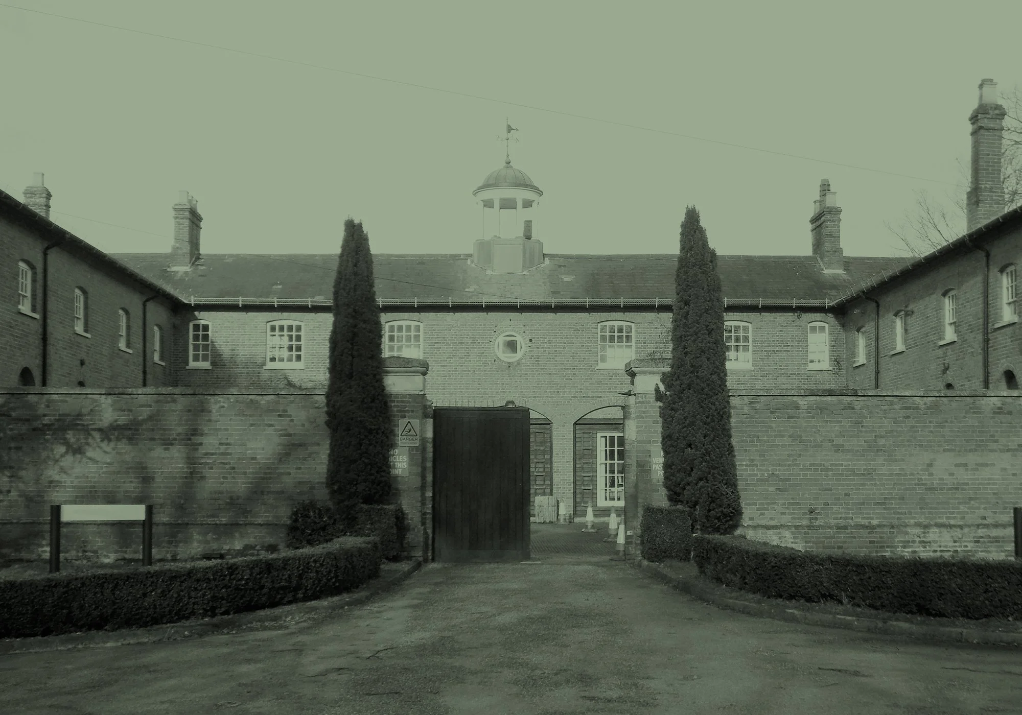 A brick building with a central tower, surrounded by a fenced courtyard with tall cypress trees, and a paved driveway leading to a gate.