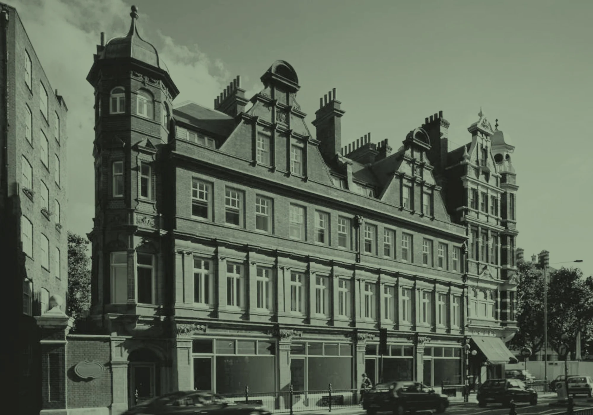 Black and white photo of a historic multi-story building with ornate rooflines and turret-like towers, located on a city street with cars parked in front.