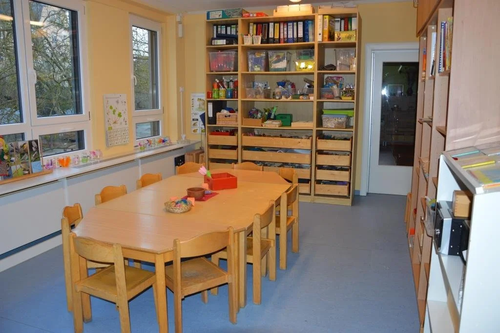 Kreativraum mit einem großen Holz-Tisch und mehreren Stühlen, Regal mit Bastelmaterialien und Spielsachen, Fenster mit Blick nach draußen, hellgelbe Wände, Tür auf der rechten Seite.