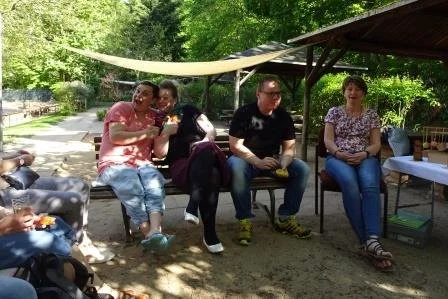Vier Menschen sitzen im Schatten unter einem Zeltdach im Freien, während sie sich unterhalten und Essen genießen.