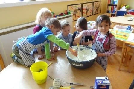 Kinder in einer Kindergartenklasse, die zusammen Teig in einer großen Schüssel mischen, während sie um einen Tisch stehen.