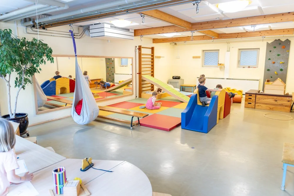 Ein Kindergartenraum mit bunten Spielmatten, Kletterwänden, einer Schaukel und Spielzeug, in dem Kinder spielen.