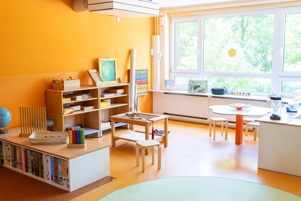 Ein heller, kinderfreundlicher Raum mit orangefarbenen Wänden, großen Fenstern und verschiedenen Spiel- und Lernspielen auf Tischen und Regalen.