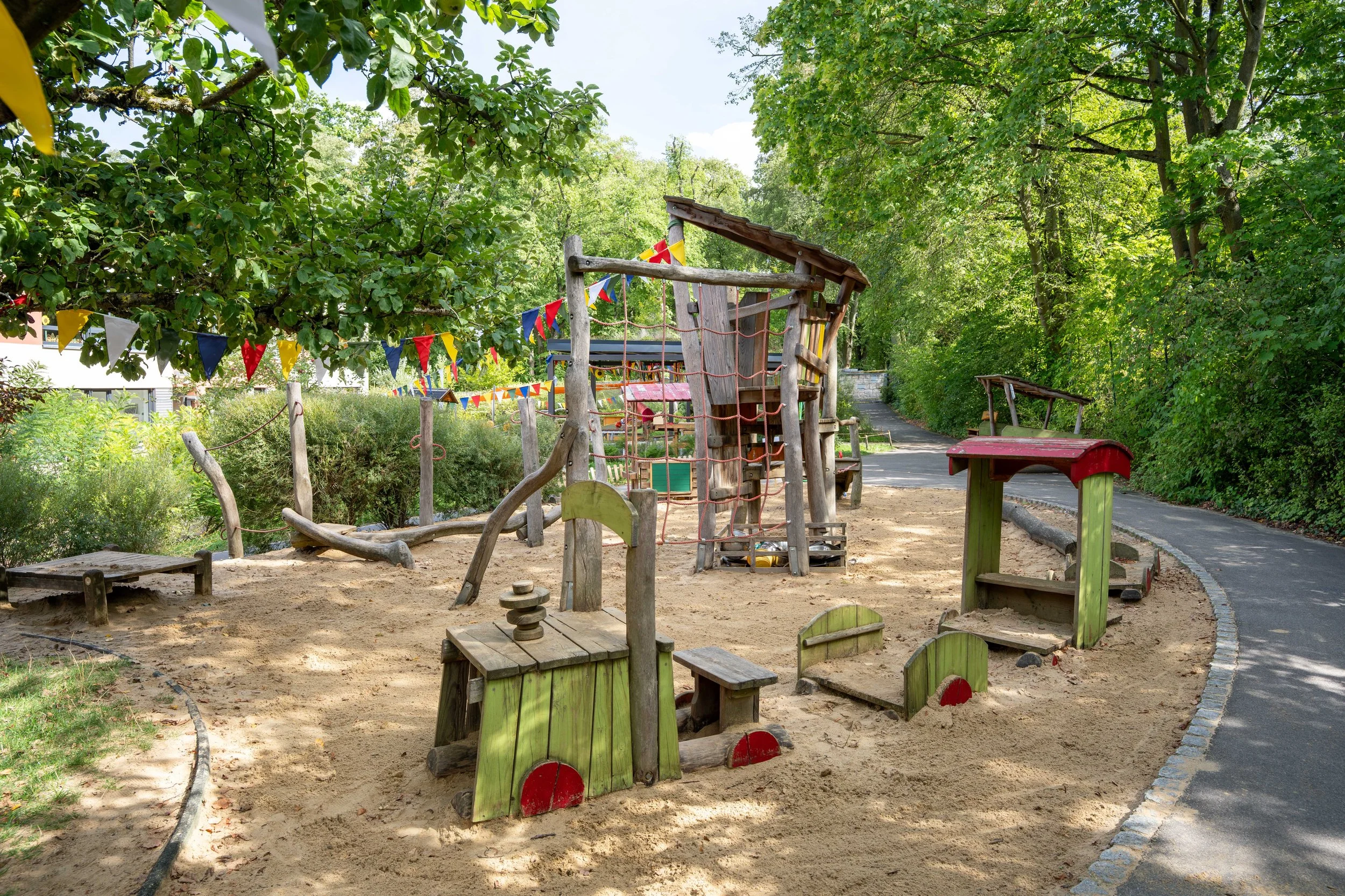 Ein Spielplatz mit Klettergerüst, Sandfläche, bunten Fahnen, umgeben von Bäumen und einer asphaltierten Straße im Hintergrund.