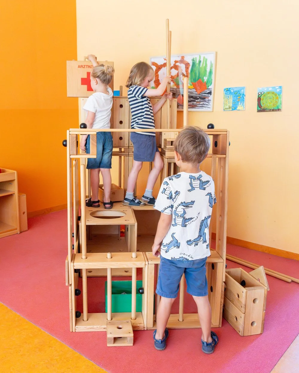 Vier Kinder spielen mit einem Holz-Klettergerüst in einem Spielzimmer mit bunten Wandbildern.