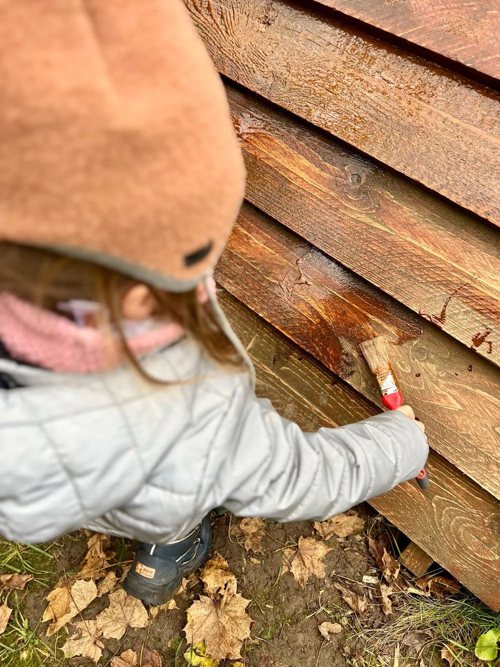 Ein Kind streicht mit einem Pinsel braunen Holzschutz auf eine hölzerne Zaunlatte.