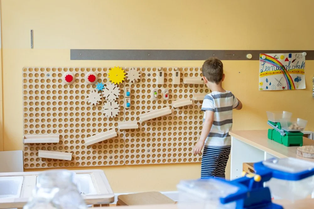 Ein Junge steht vor einer Wandtafel mit einem Holz-Puzzle, das in einer Kindertagesstätte für Lernspiele genutzt wird, mit bunten Bildern und Materialien im Vordergrund.