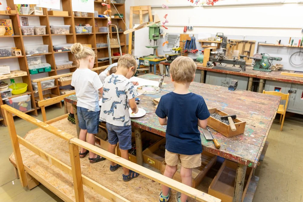 Kinder in einem Holzwerkstattraum beim Basteln und Arbeiten mit Holz am Tisch, umgeben von Werkzeuge und Holzmaterialien.