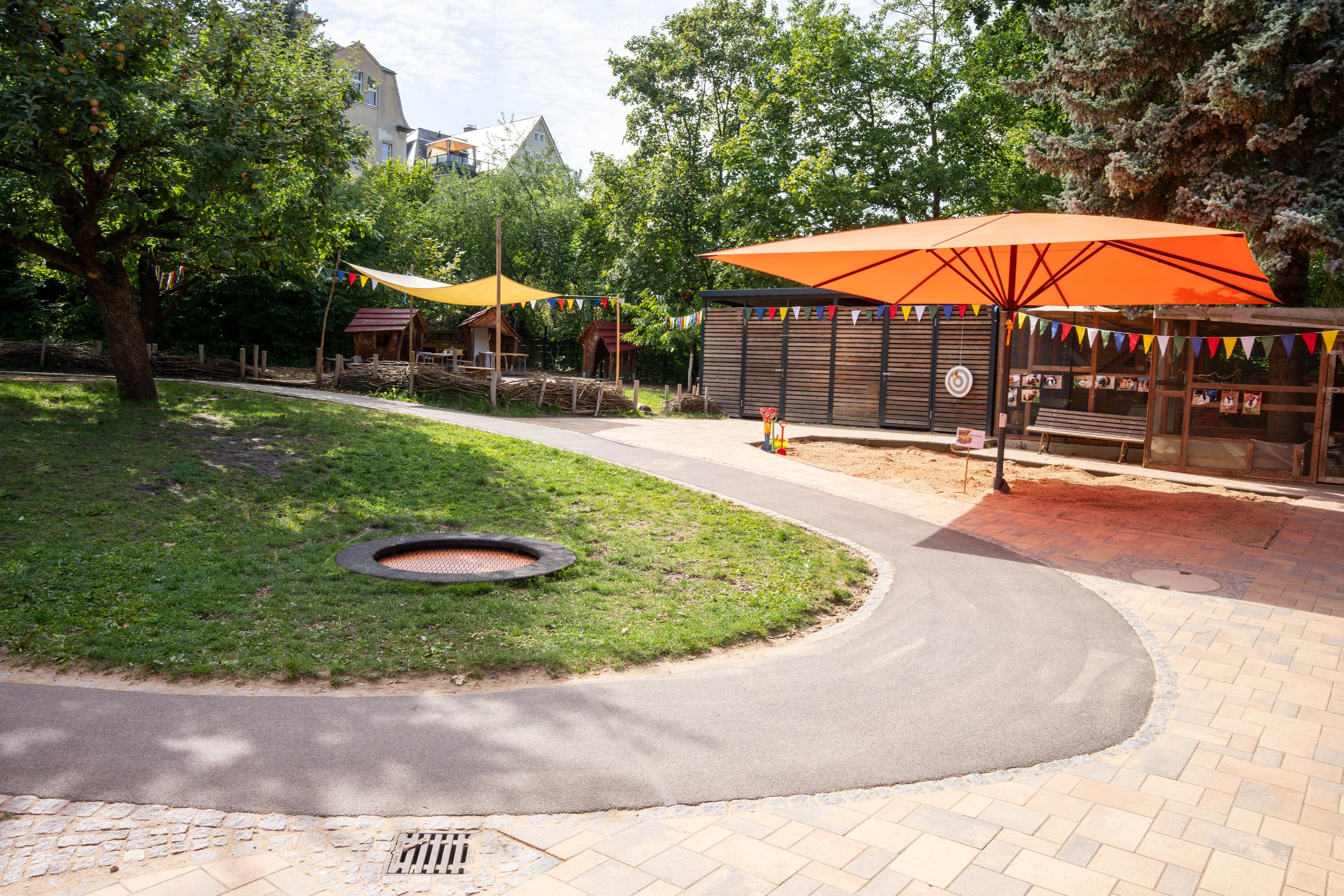 Ein Spielbereich im Freien mit Bäumen und buntem Sonnenschutz, inklusive Trampolin, Sandkasten, Zielscheibe und Wasserpistolen.