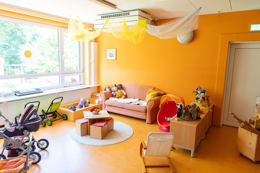 Ein Spielzimmer mit orangefarbenen Wänden, einem rosa Sofa, Spielzeugen, Puppen und Kinderwagen, Sonnenlicht durch große Fenster, und Dekorationen wie zarten Stoffbahnen an der Decke.