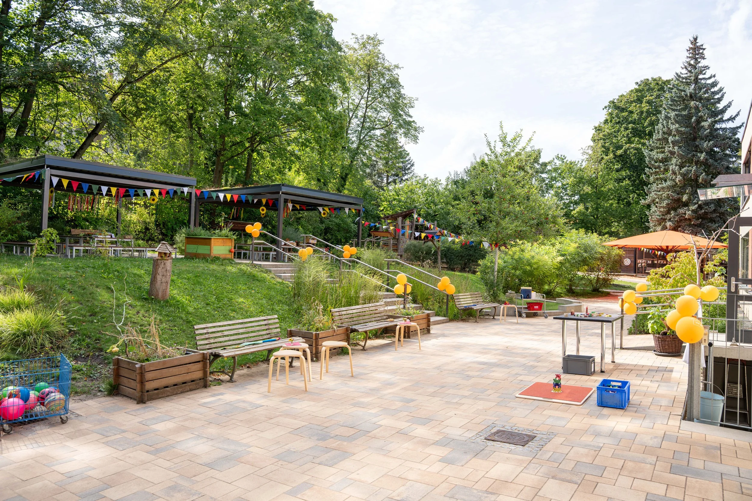 Freiluft-Spielplatz mit Bänken, bunten Flaggen und Luftballons, umgeben von Bäumen und einem Gebäude im Hintergrund.