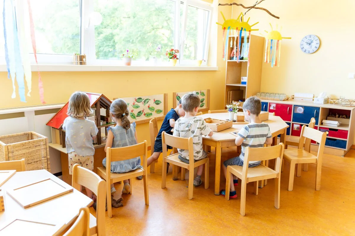 Kinder spielen und basteln in einem bunten, freundlichen Kindergartenraum mit großen Fenstern, dekorativen Papiergirlanden, einem Vogelhaus, einer Wand mit grünem Blattmuster und bunten Sonnen-Dekorationen.