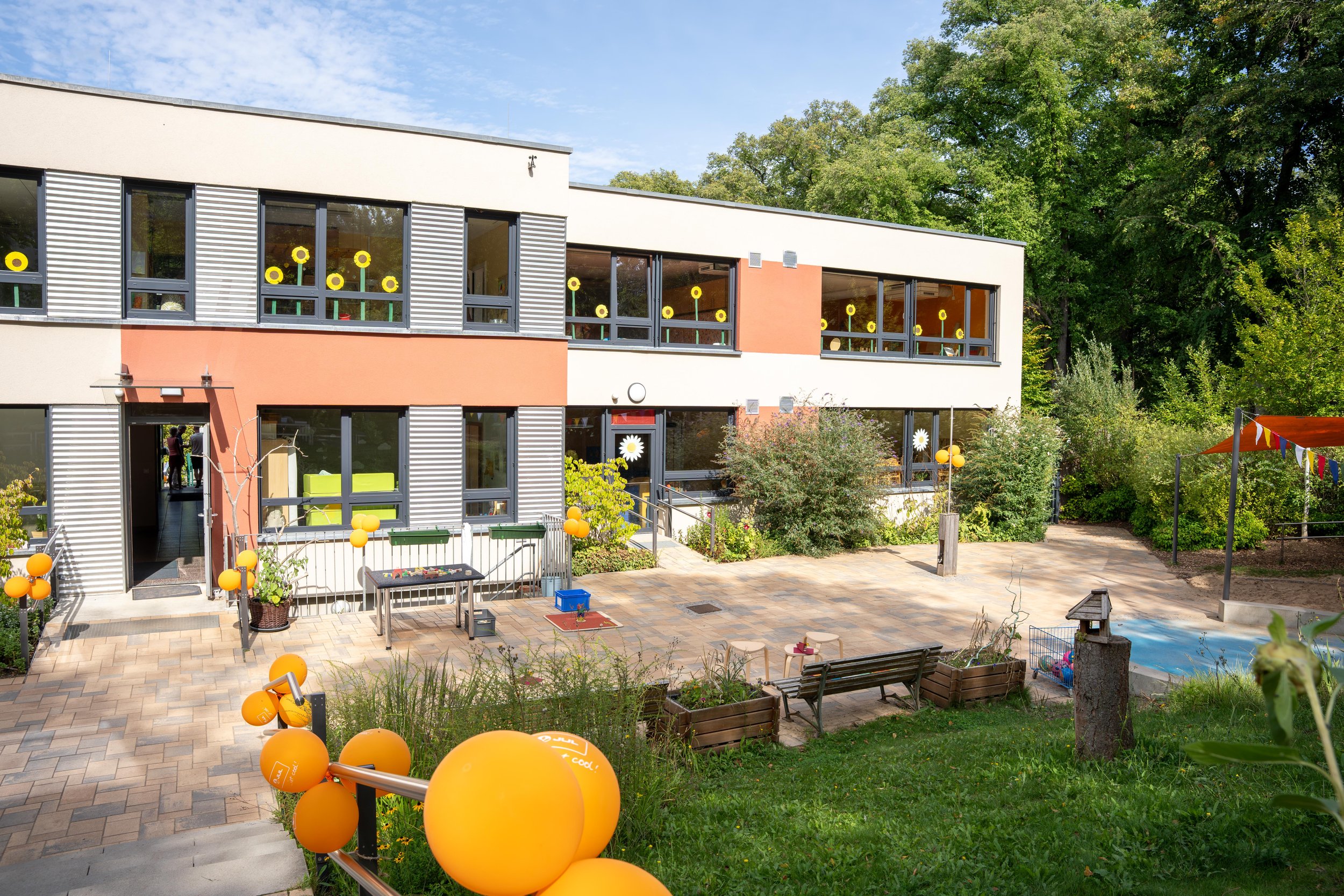 Mehrstöckiges Gebäude mit großen Fenstern und bunten Dekorationen, umgeben von grünen Bäumen, auf einem gepflasterten Innenhof mit Spielzeug, Sitzgelegenheiten und Sommerdekorationen.