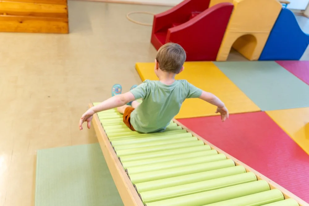 Junge auf einer grünen Rutschmatte in einem Spielzimmer mit buntem Klettergerät aus Holz in rot, gelb, blau und grün