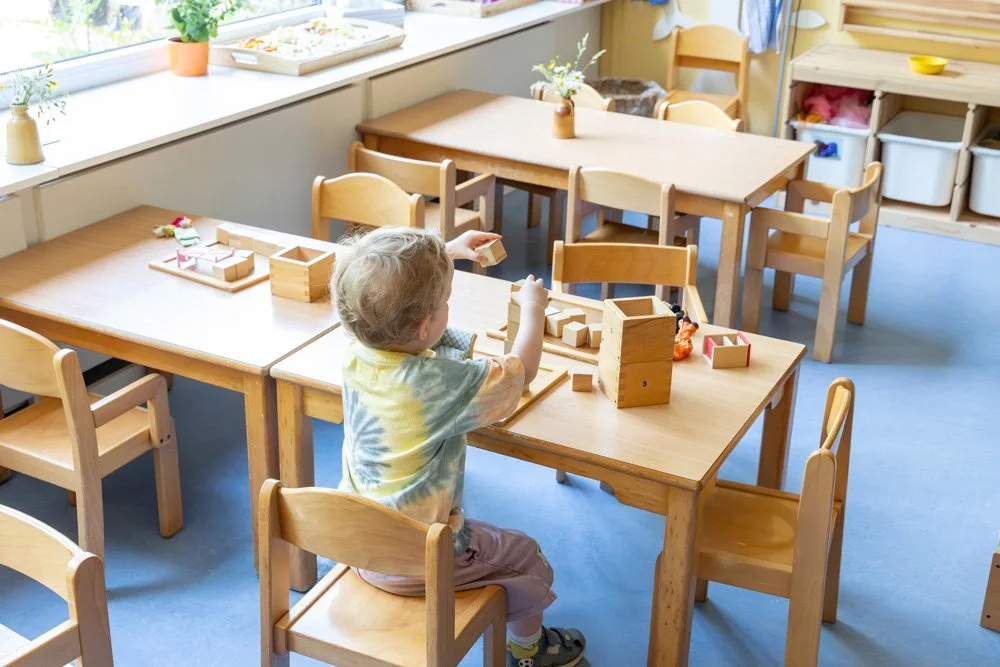 Ein Kinderzimmer mit mehreren Holztischen und Stühlen, ein Kind spielt mit Holzbausteinen, Fenster mit Pflanzen im Hintergrund.