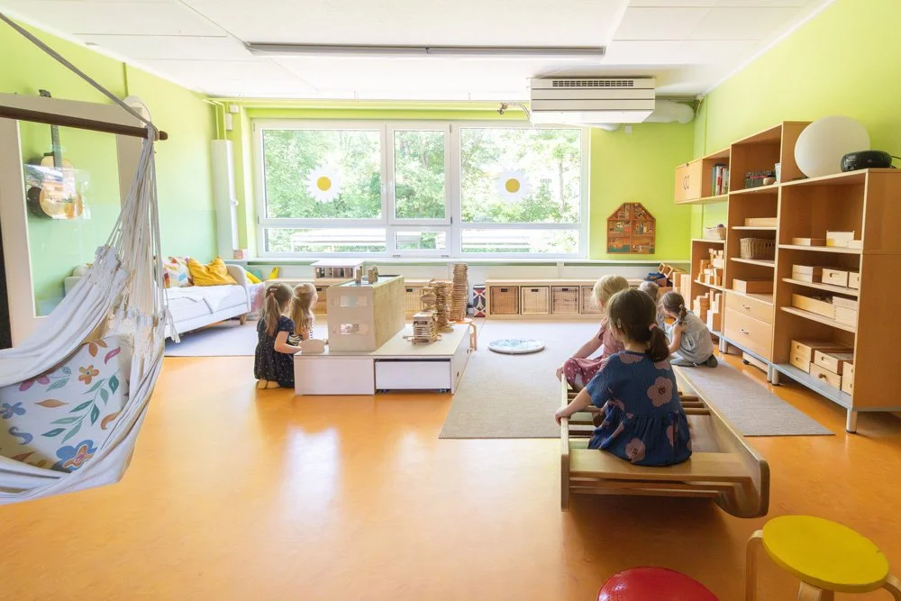 Ein hell erleuchteter Kindergartenraum mit vier Kindern, die am Boden sitzen, und einer gemütlichen Sitzecke mit Kissen. Der Raum hat große Fenster mit grüner Aussicht, Regale mit Spielzeug und Büchern, sowie eine Hängematte an der linken Seite.