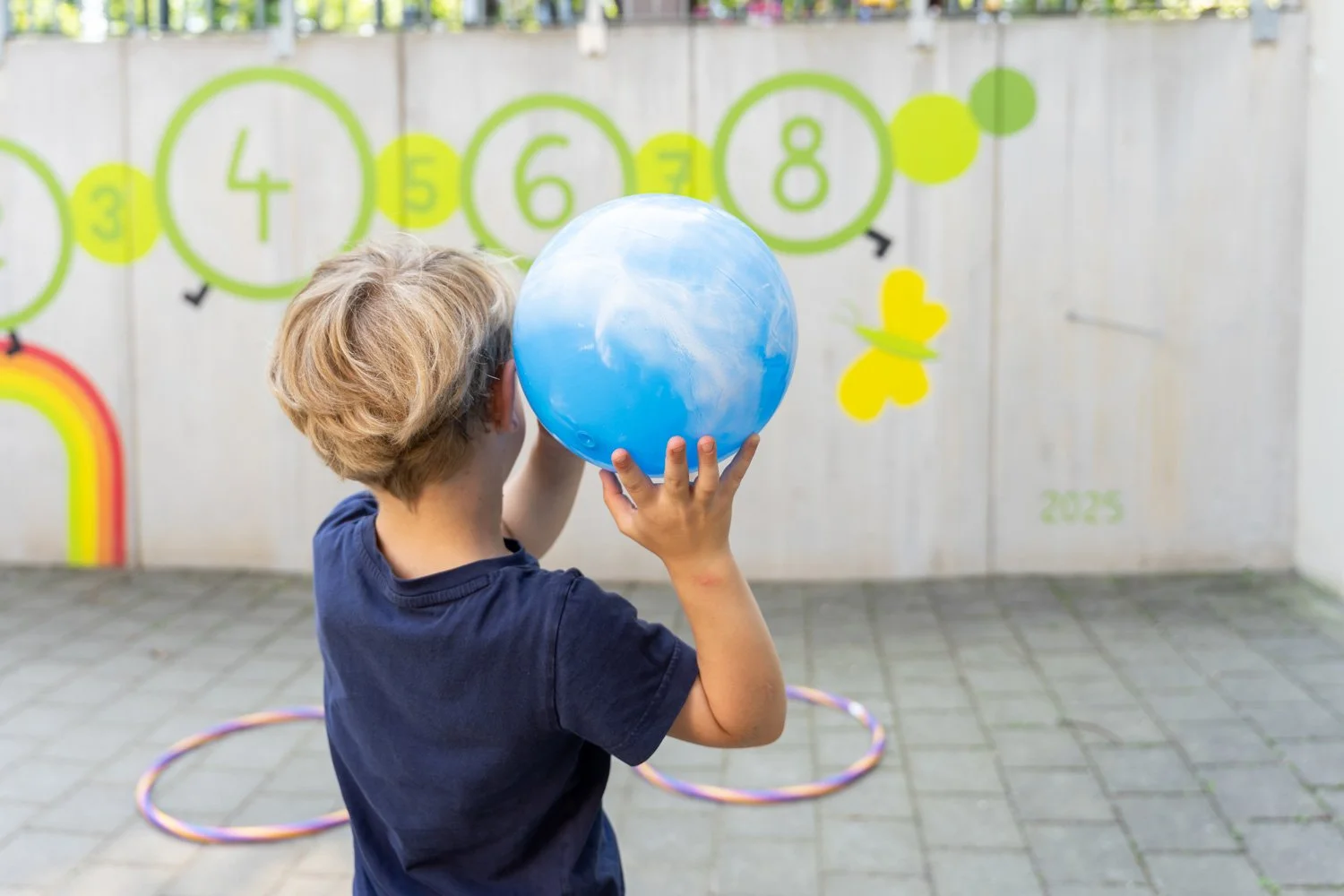Junge spielt mit einem blauen Ball im Außenbereich, im Hintergrund eine Wand mit Lernzeichen und Käfern, Hula-Hoop-Reifen auf dem Boden.