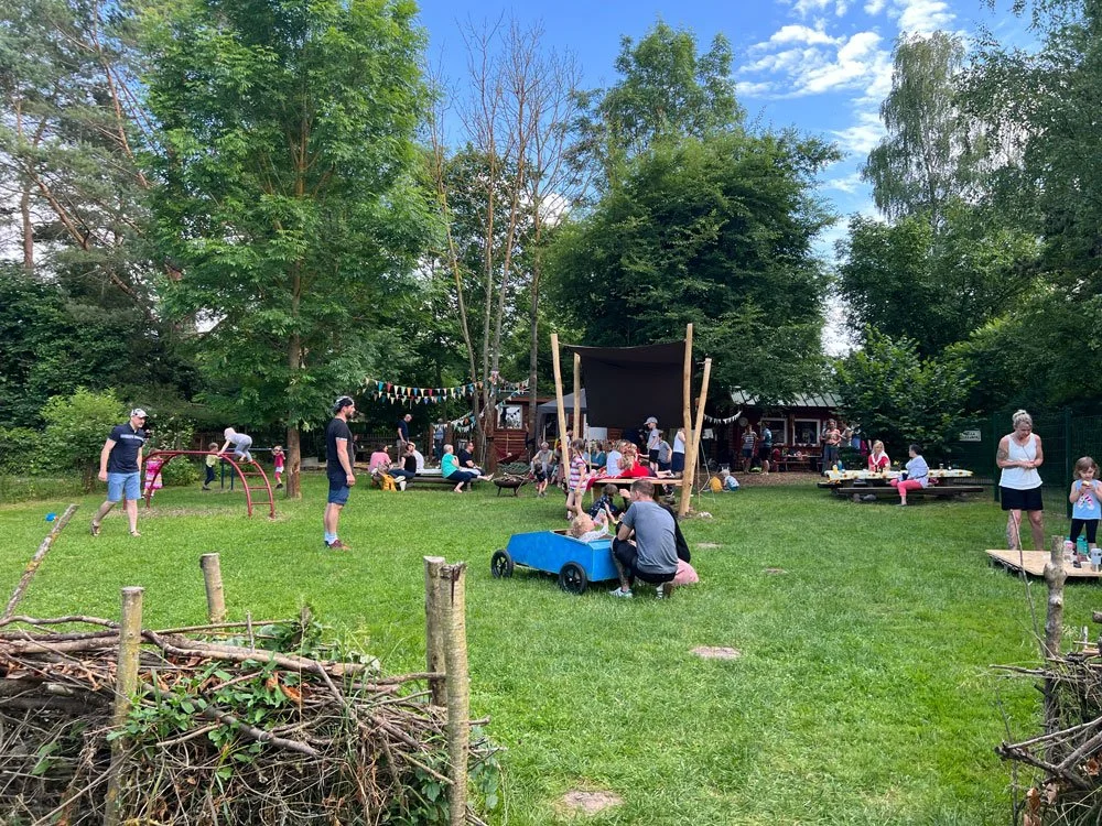 Grüner Garten mit vielen Kindern und Erwachsenen, aktive Spiele, Sitzgruppen, Bäume, blauer Himmel, festliche Dekoration, Holzwege