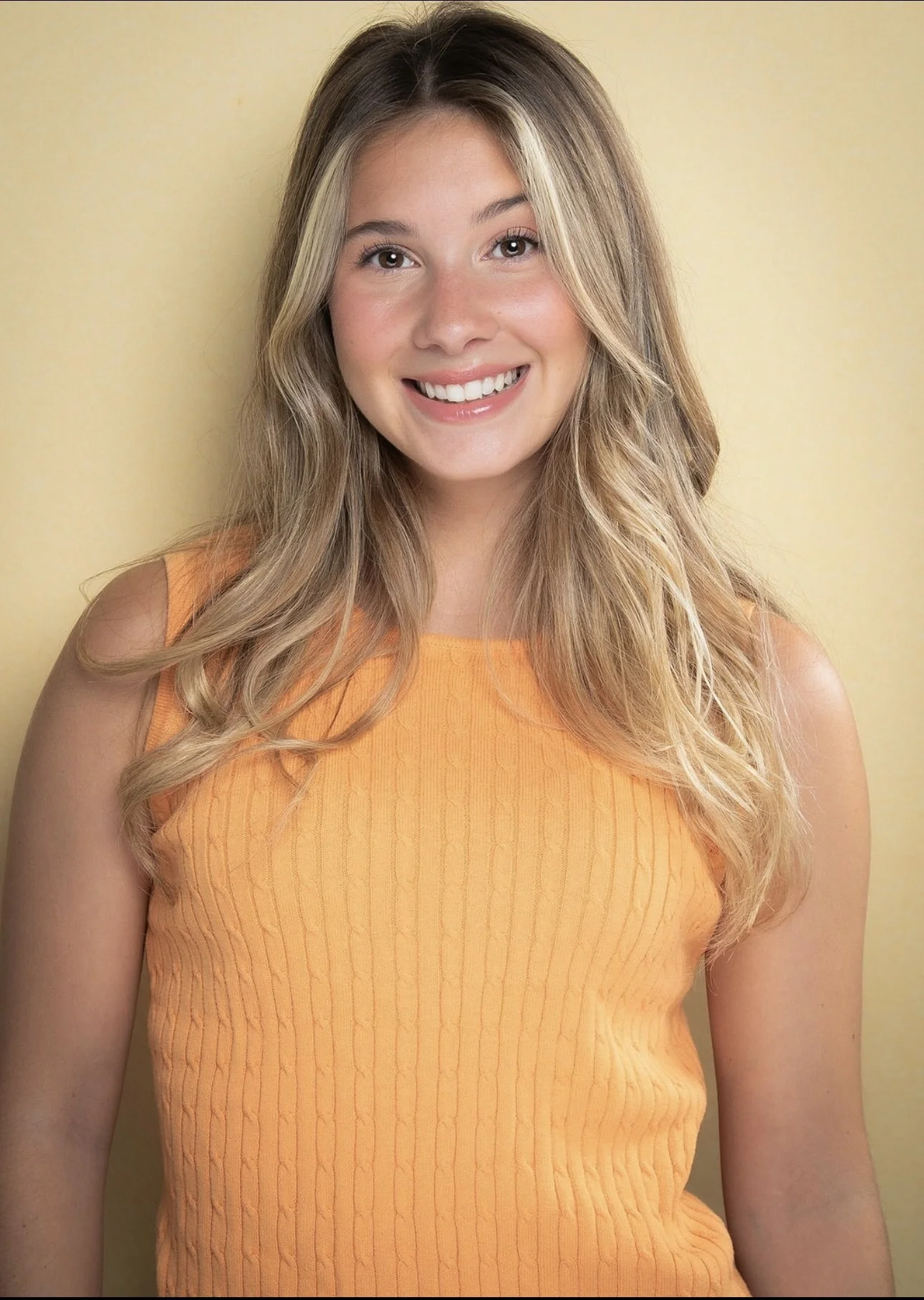 A young woman with long, wavy blonde hair, smiling, wearing an orange sleeveless top, standing against a yellow background.