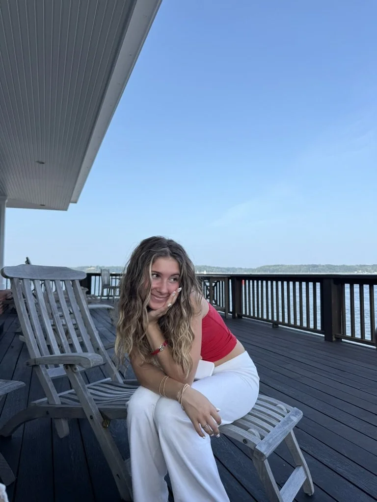 A young woman with wavy hair wearing a red shirt and white pants, sitting on a wooden deck overlooking a body of water with a clear blue sky.
