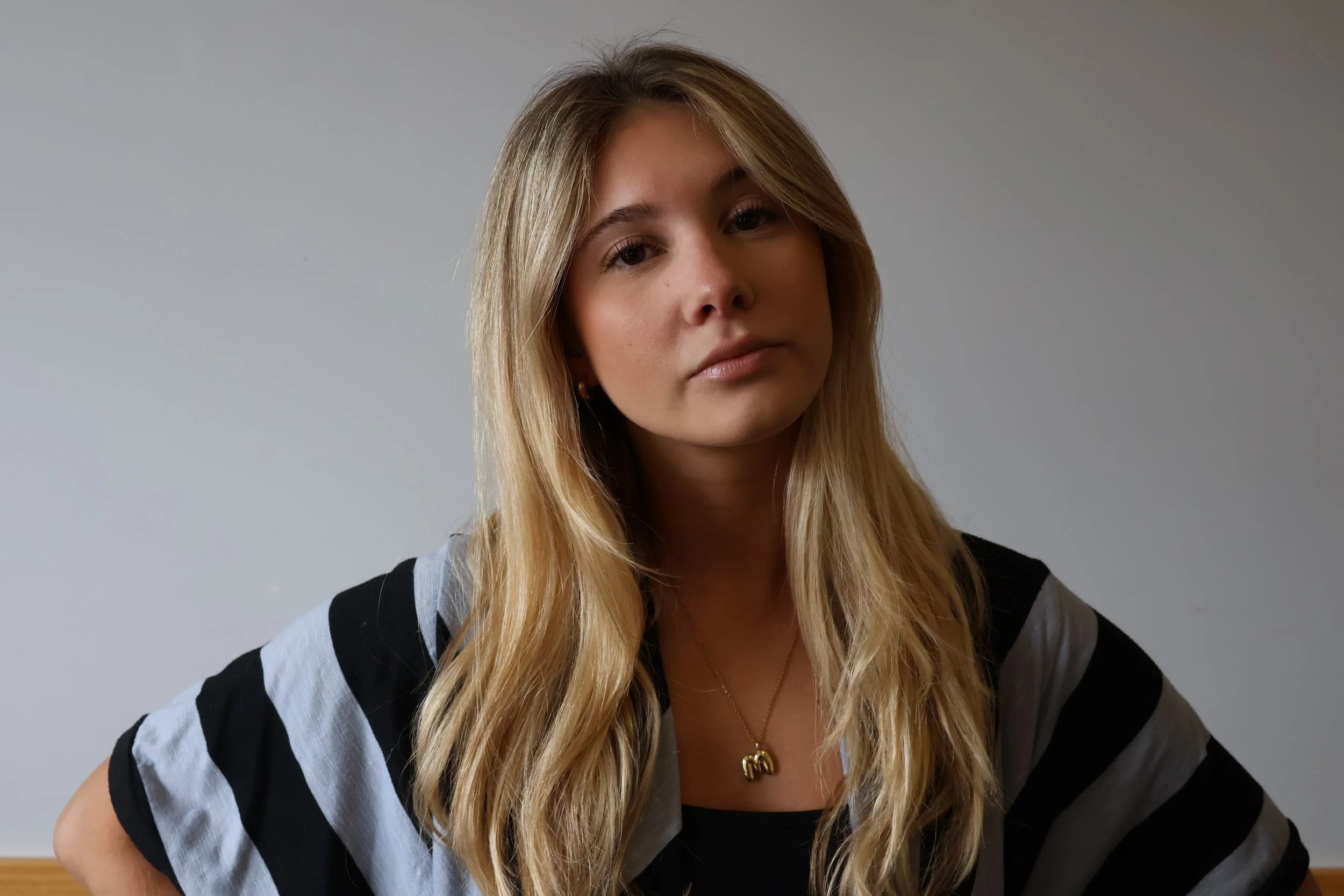 A young woman with long blonde hair and a serious expression poses against a plain gray background. She is wearing a black and white striped shirt and a gold necklace with a pendant.
