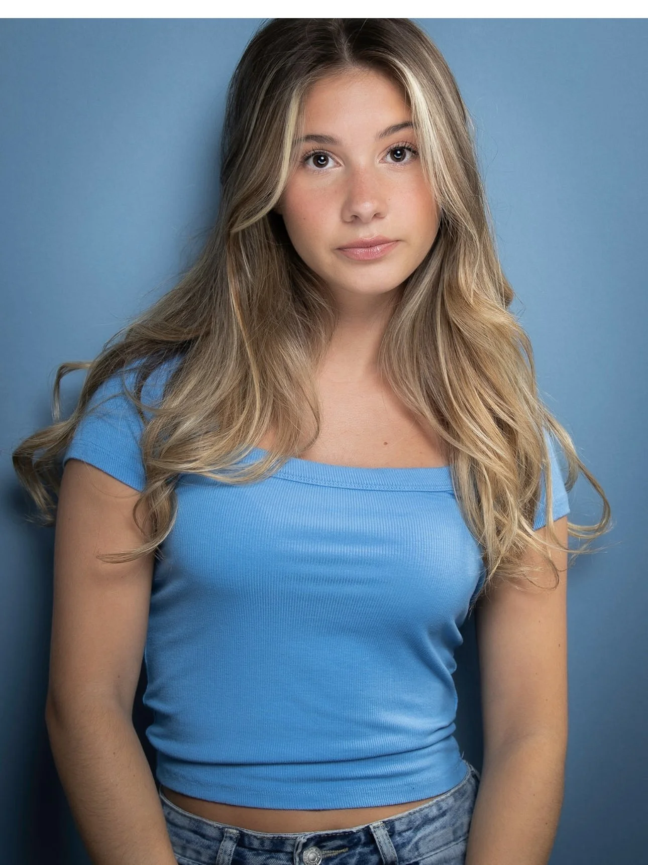 A young woman with long, wavy blonde hair wearing a fitted blue T-shirt and denim jeans stands against a blue background, looking directly at the camera with a neutral expression.