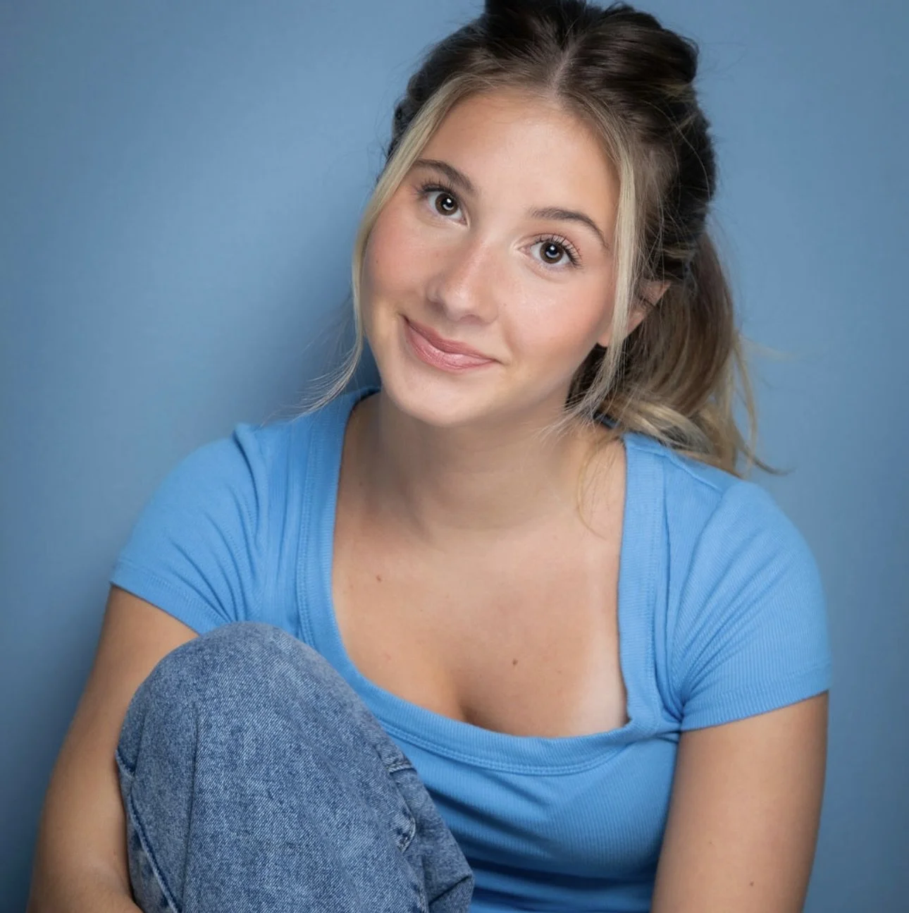 A young woman with blonde and brown hair in a ponytail, wearing a blue t-shirt, sitting against a blue wall, smiling softly at the camera.