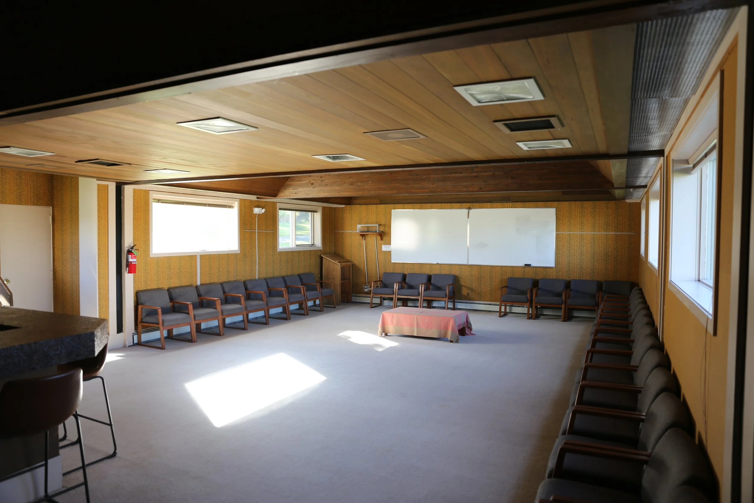 Empty meeting room with chairs arranged along the walls, a table covered with a pink tablecloth, and windows letting in natural light.