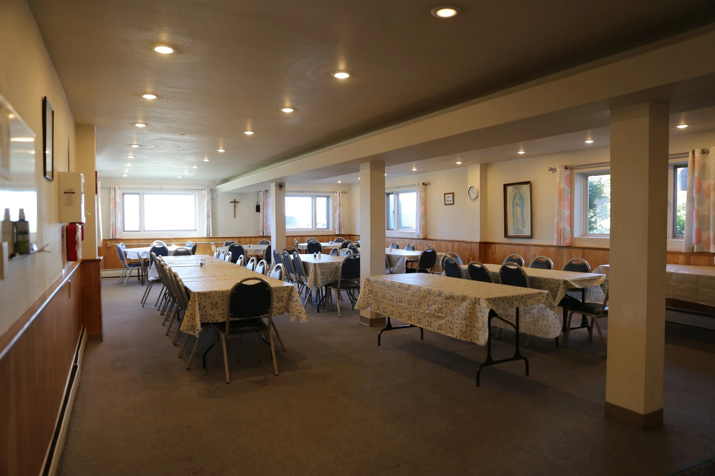 A dining hall with multiple rectangular tables covered in floral tablecloths and chairs around them, illuminated by ceiling lights, with windows on the back wall and religious decorations like a crucifix and a picture of the Virgin Mary.