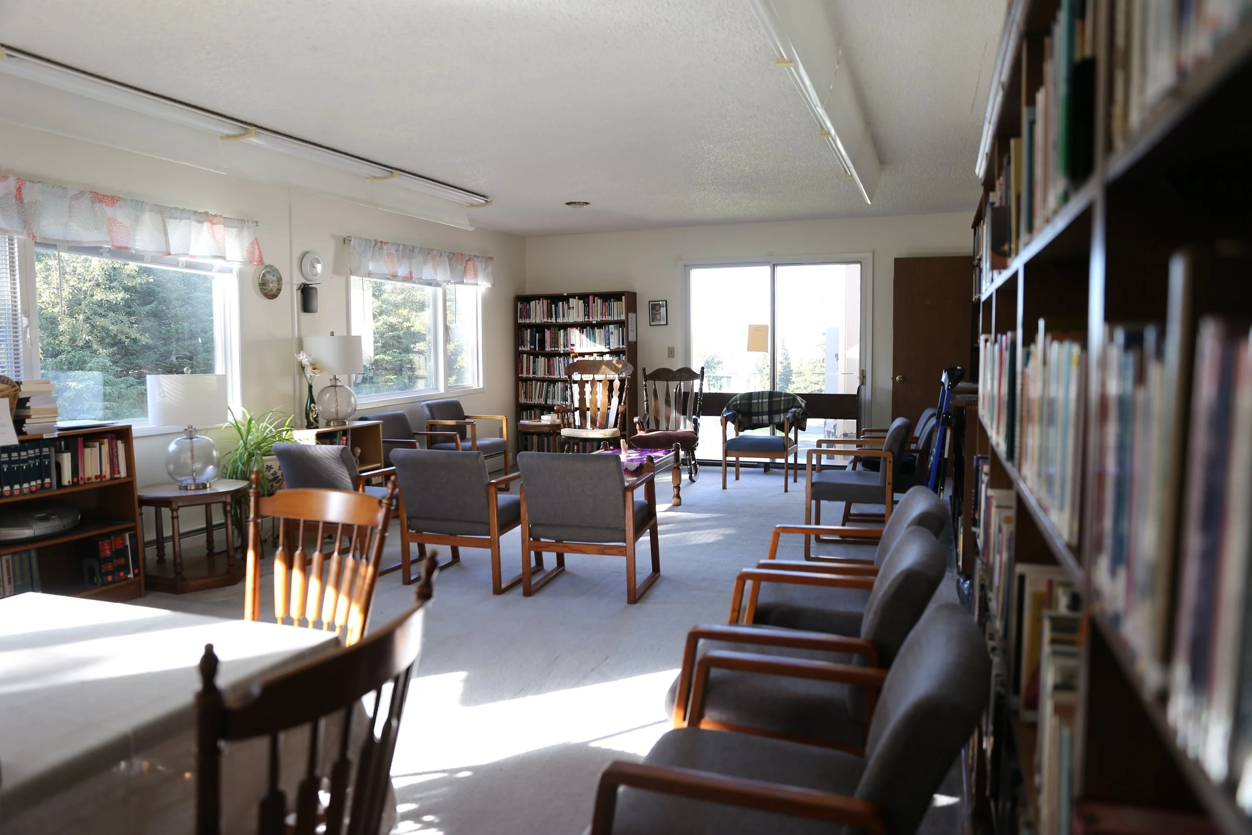 A cozy library or reading room with bookshelves filled with books on the right, multiple chairs arranged in groups for seating, large windows let in natural light, and there is a door leading outside. Sunlight casts shadows on the floor.