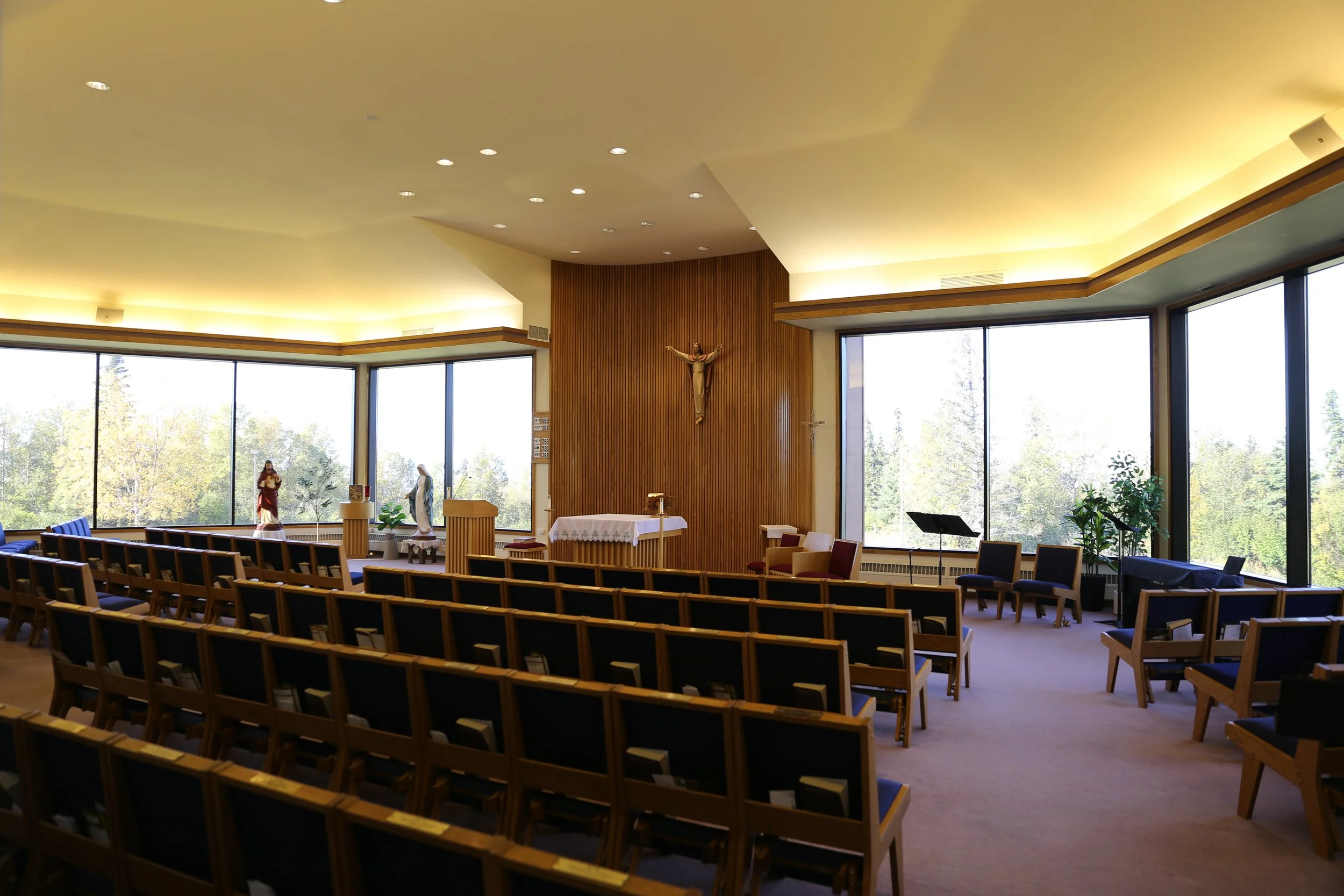 Interior of a church sanctuary with wooden pews, religious statues, and large windows showing trees outside, with soft lighting and a wooden altar area.