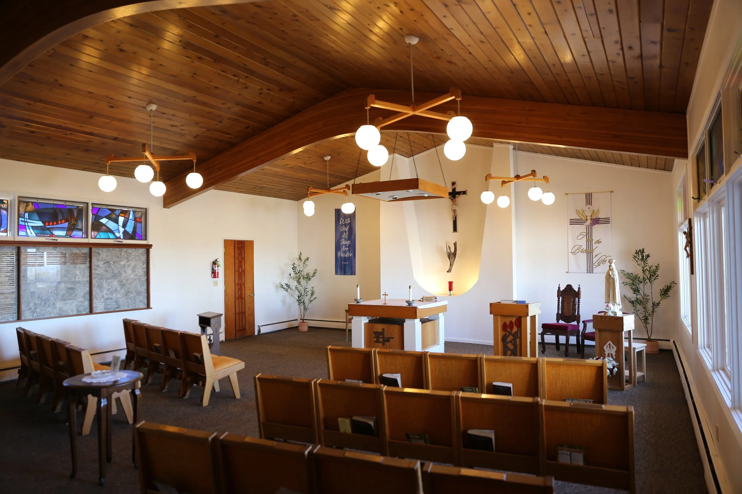 Interior of a church with wooden ceiling, chandeliers, stained glass windows, altar, and chairs arranged in rows.