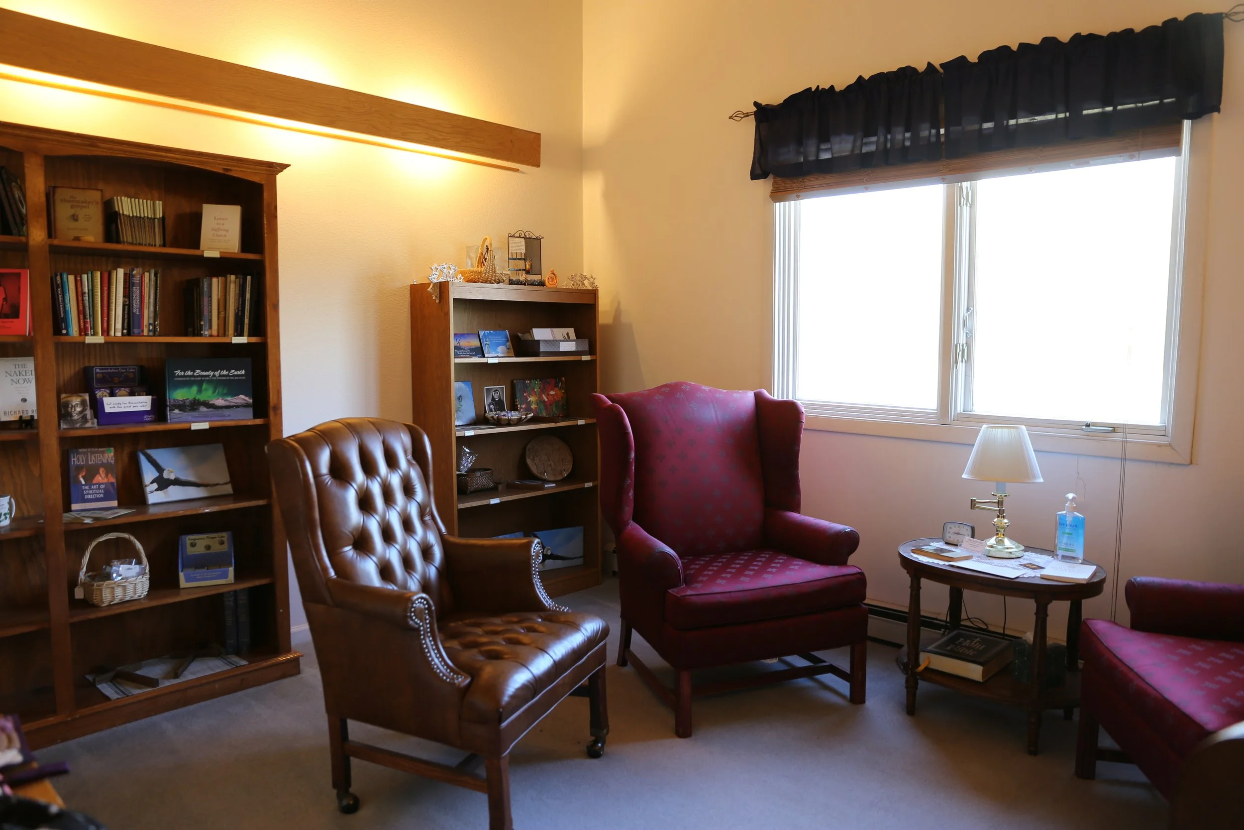 A cozy sitting room with red upholstered armchairs, a wooden bookshelf filled with books, a small table with a lamp, and a large window with black curtains letting in sunlight.