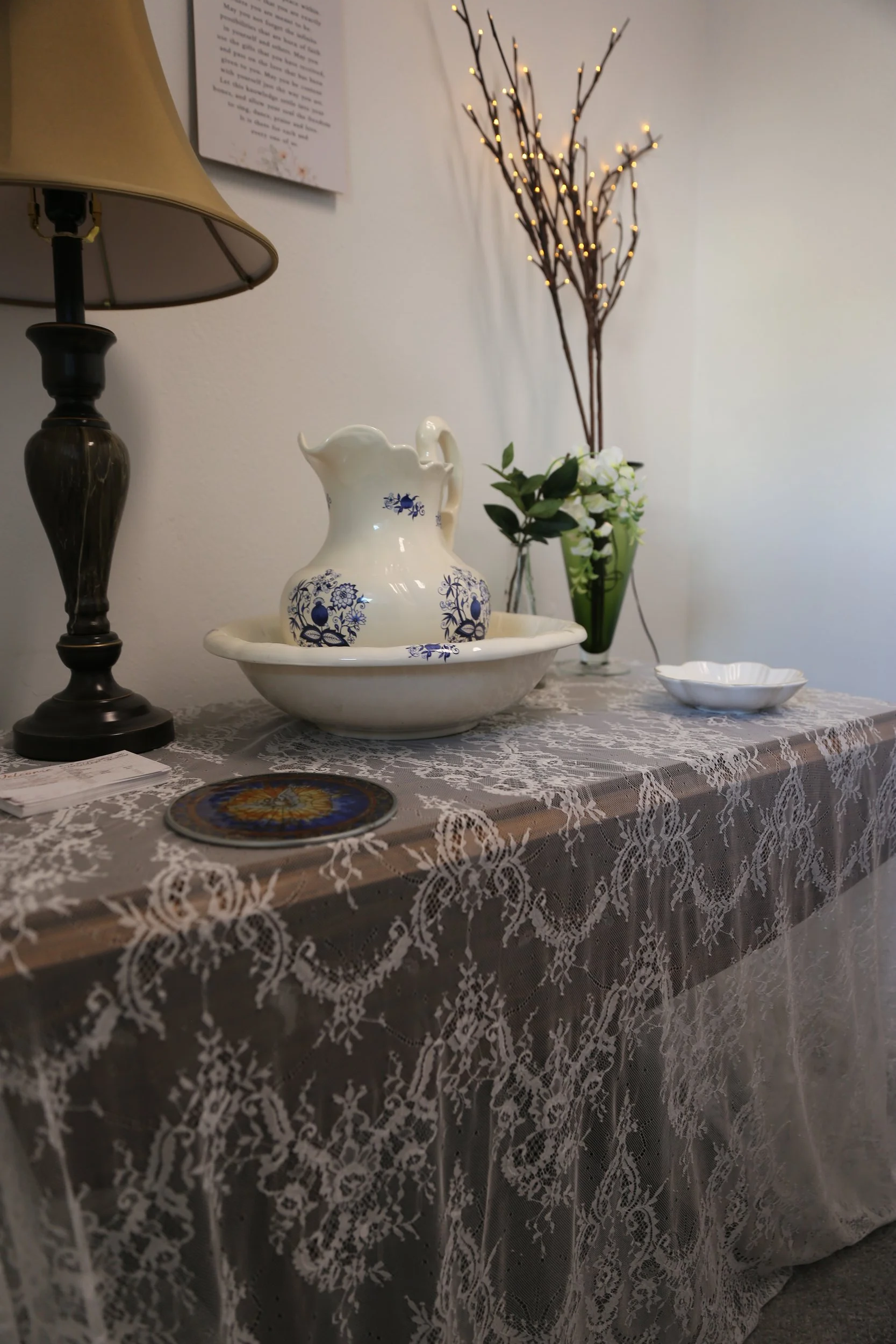 A table with a lace tablecloth, a ceramic pitcher with blue floral patterns, a green vase with white flowers, a decorative plate, a lamp, and a framed piece of paper on the wall.