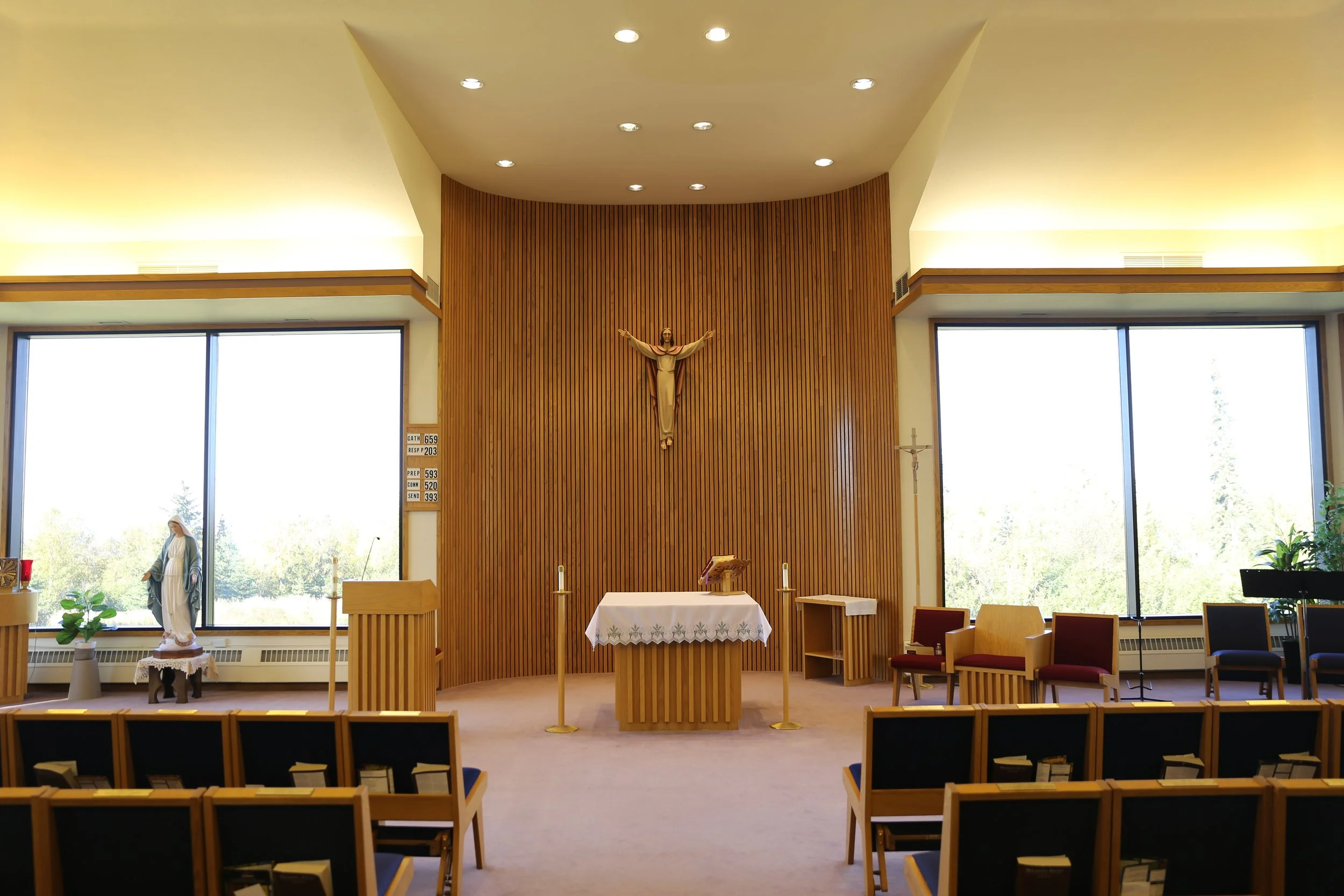 Interior of a Catholic church with altar, crucifix, and religious statues, illuminated by large windows and ceiling lights.