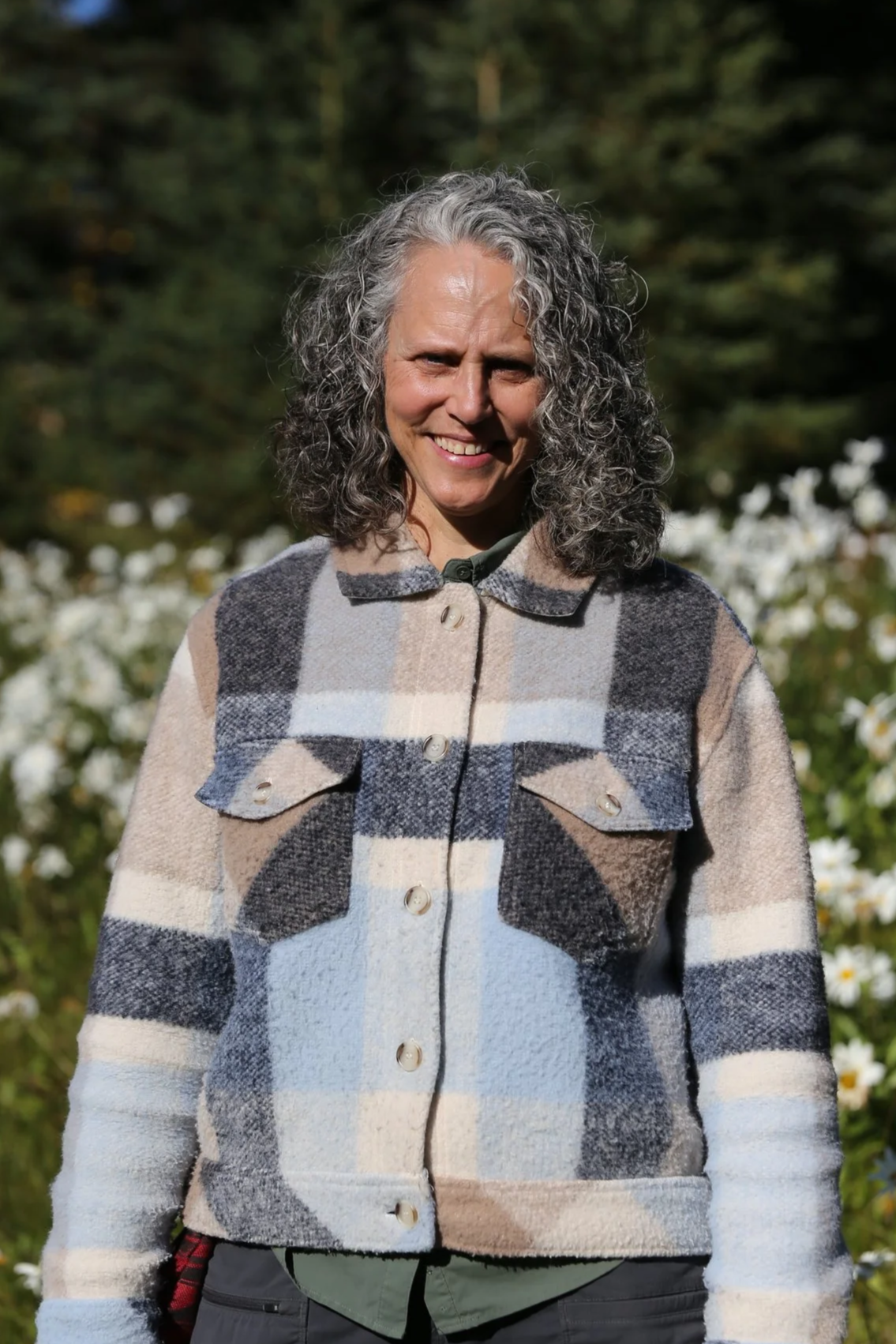 A smiling woman with curly gray hair wearing a plaid fleece jacket outdoors with trees and white flowers in the background.