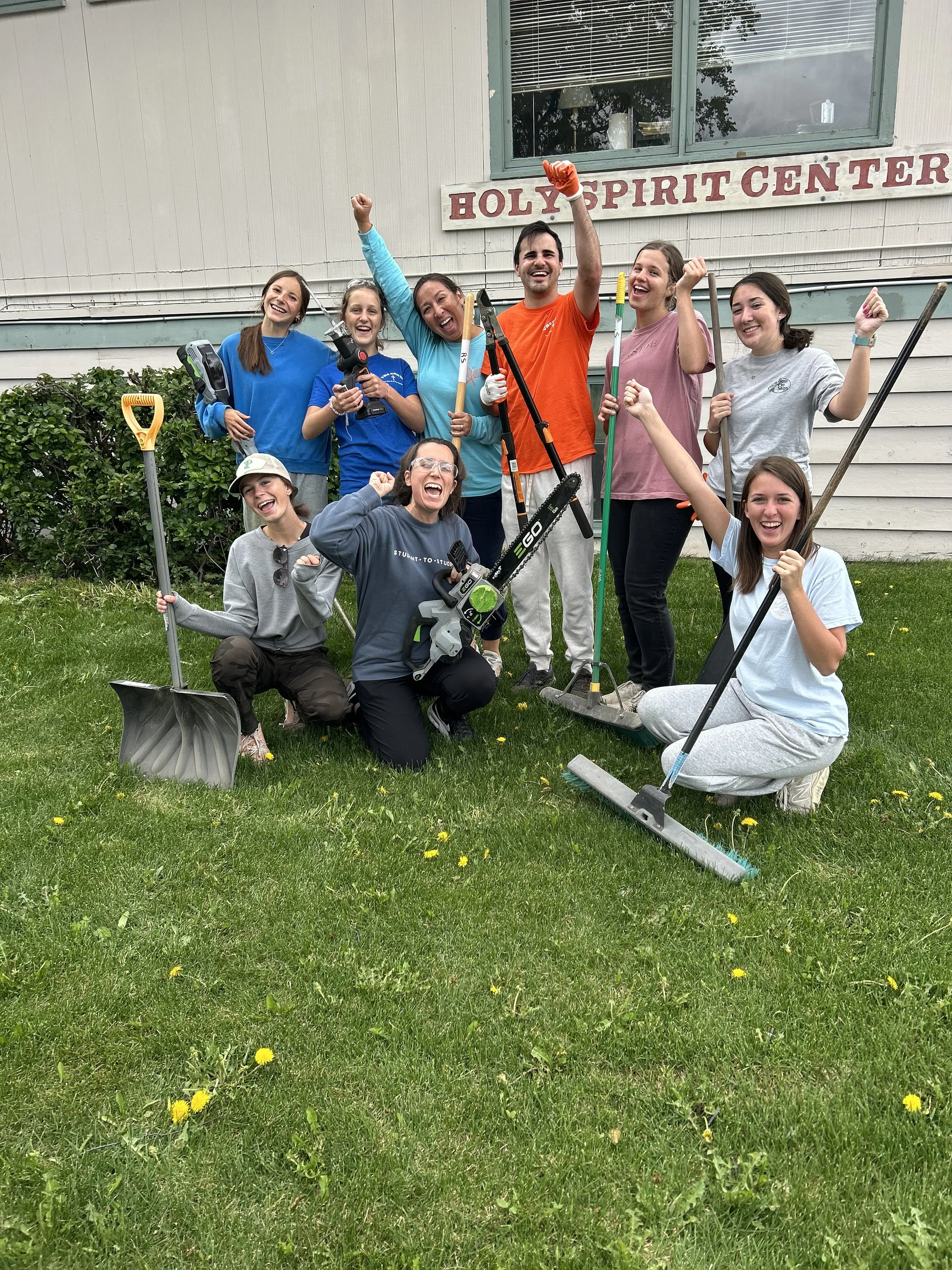 Group of people outdoors, celebrating the completion of community service at a church named Holy Spirit Center, holding gardening tools, smiling, and posing on a lawn.