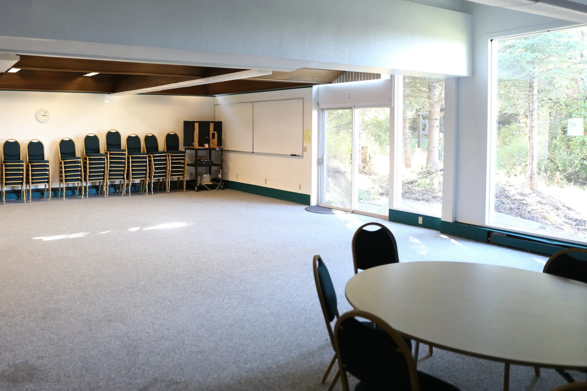 Empty room with stacked chairs along the wall, round tables with black chairs, large windows showing trees outside, and a whiteboard with a clock on the wall.