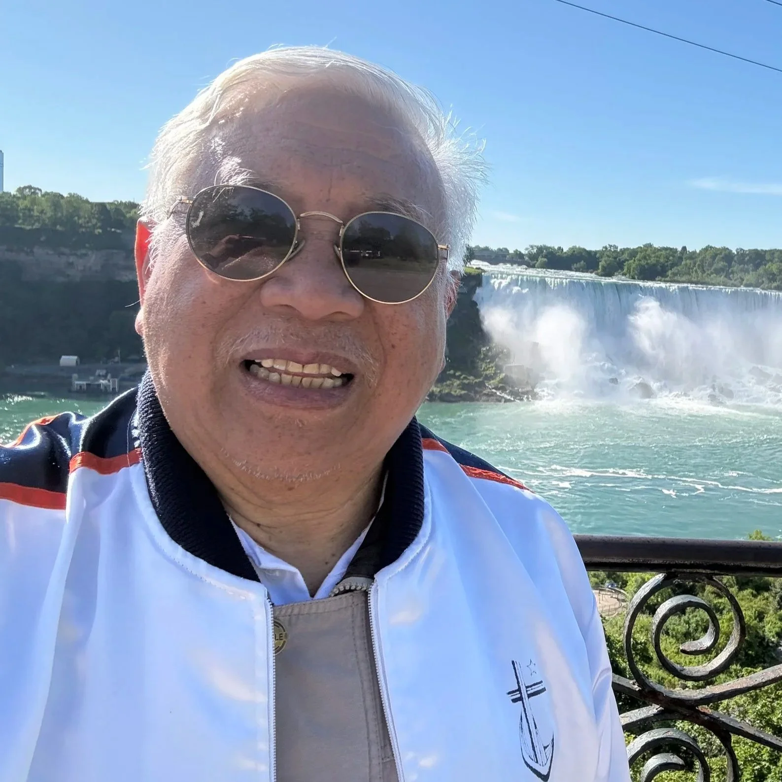 A man with white hair wearing sunglasses and a white jacket taking a selfie with Niagara Falls in the background on a sunny day.