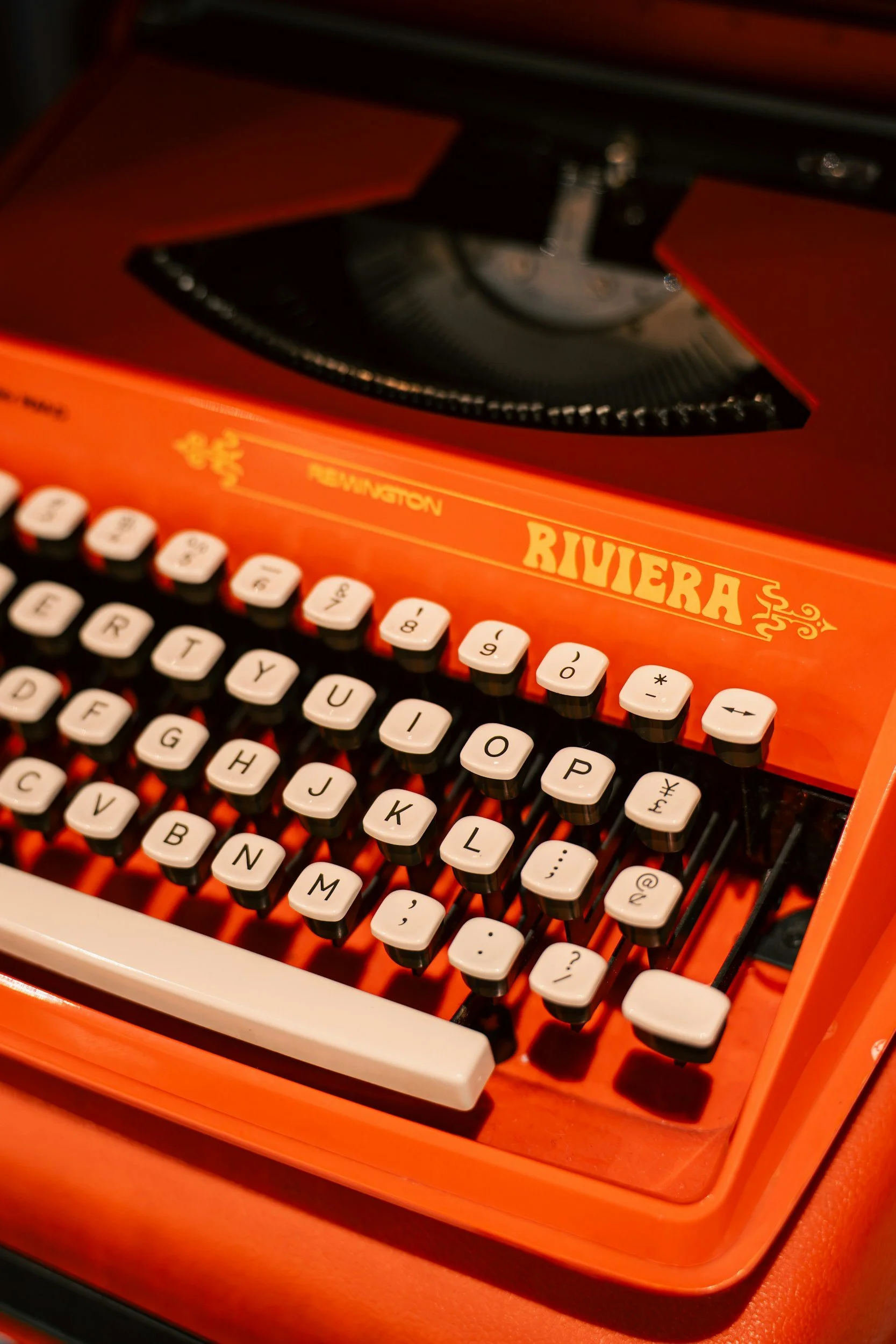 Close-up of a vintage orange typewriter with white keys, the brand name 'Riviera' on the upper front, and a black roller at the top.