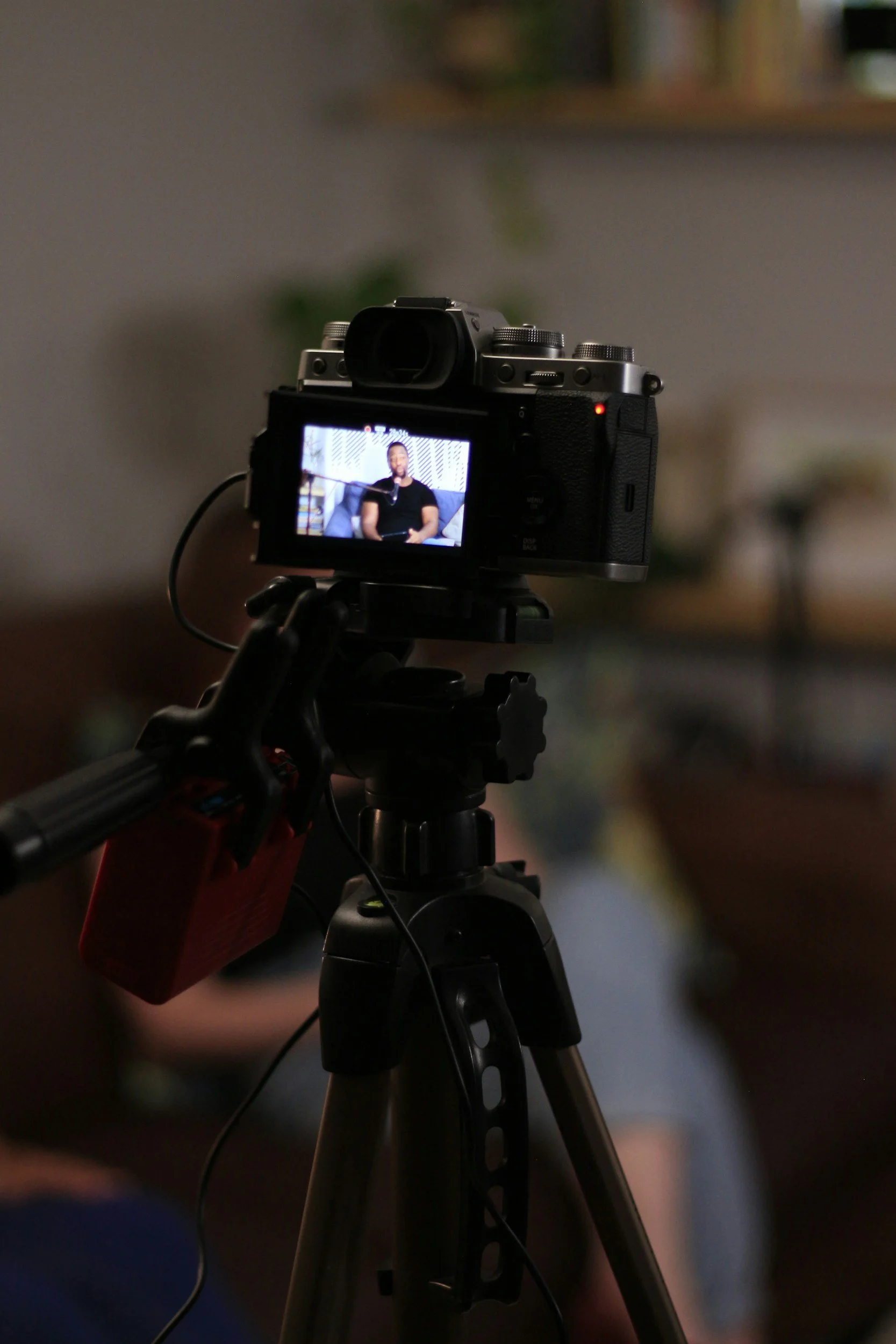 A professional camera mounted on a tripod, with a screen displaying a man sitting on a sofa in a studio environment.