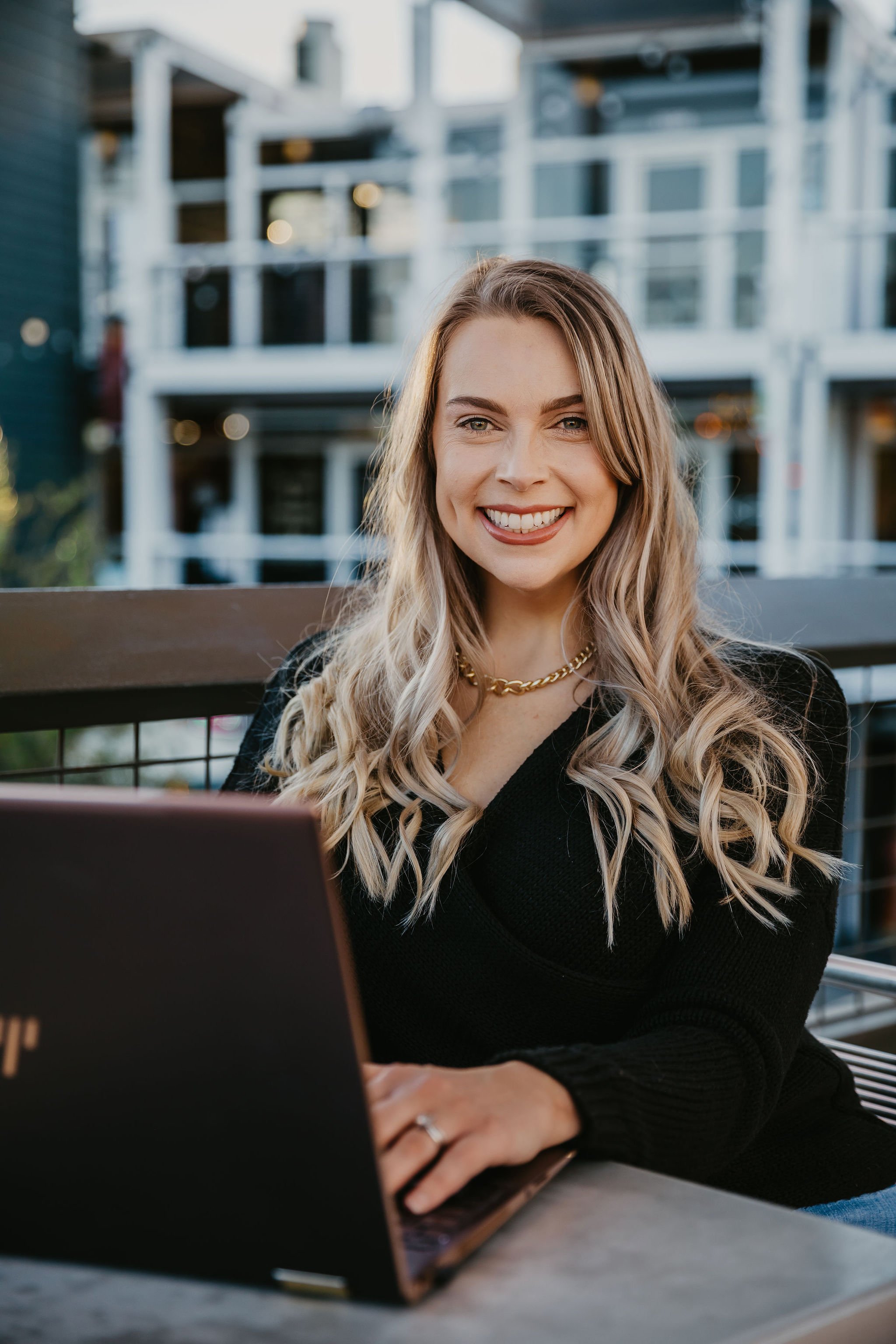 A smiling woman with blonde wavy hair, wearing a black top and a gold chain necklace, sitting outdoors at a table with a laptop, with a blurred modern apartment building in the background.