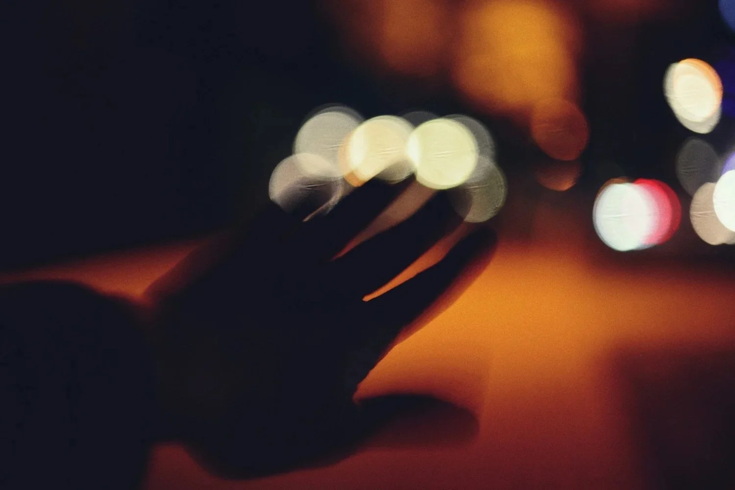 A person's hand reaching out in the dark with blurred city lights in the background, creating bokeh effects.