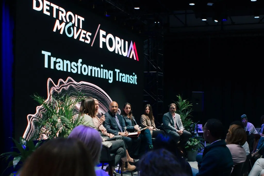 A panel of five speakers at Detroit Moves Forum discussing transforming transit, with an audience in front and decorative plants around.
