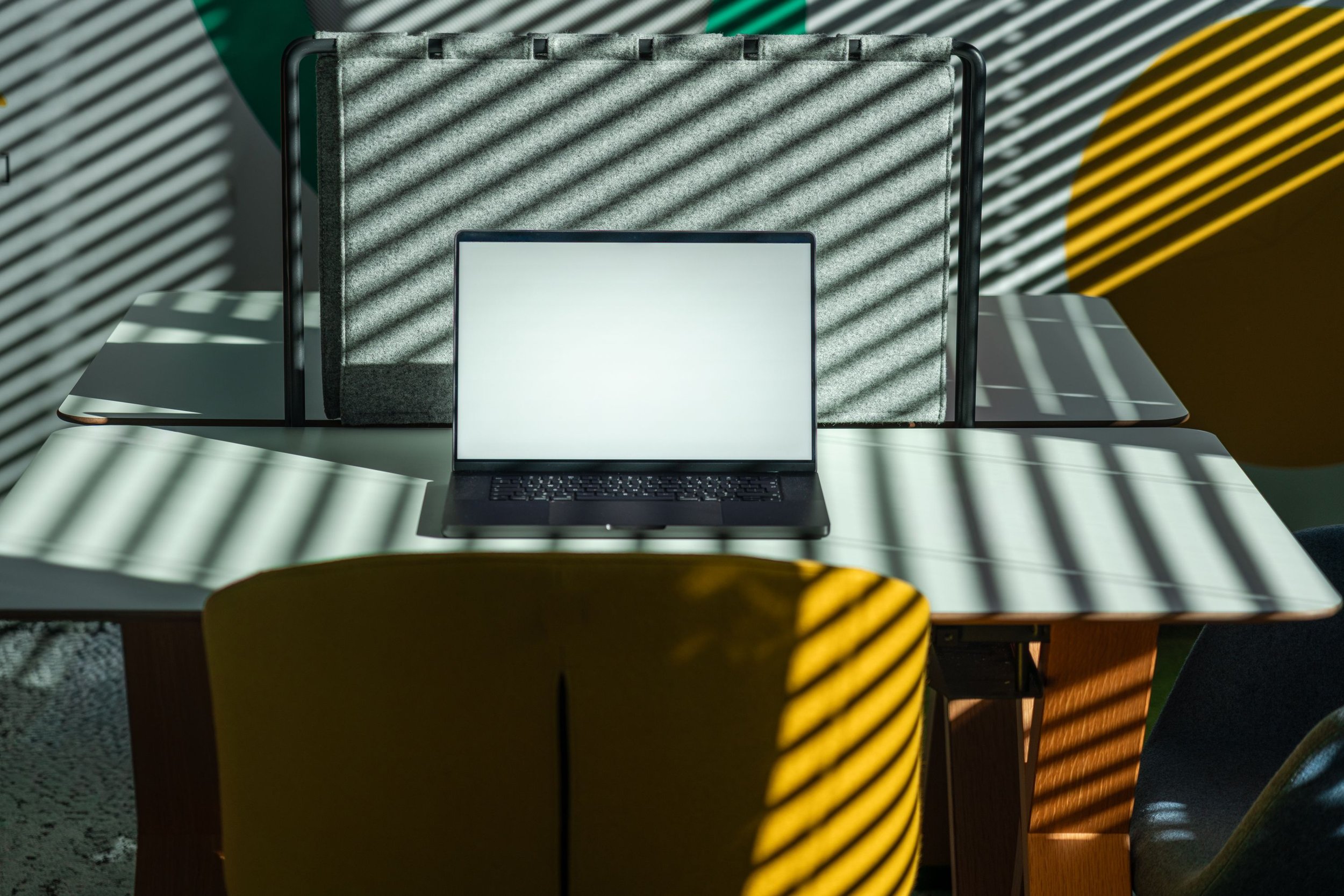 Laptop on a white table with sunlight and shadows cast from window blinds, yellow and grey chairs, and a textured grey wall in the background.