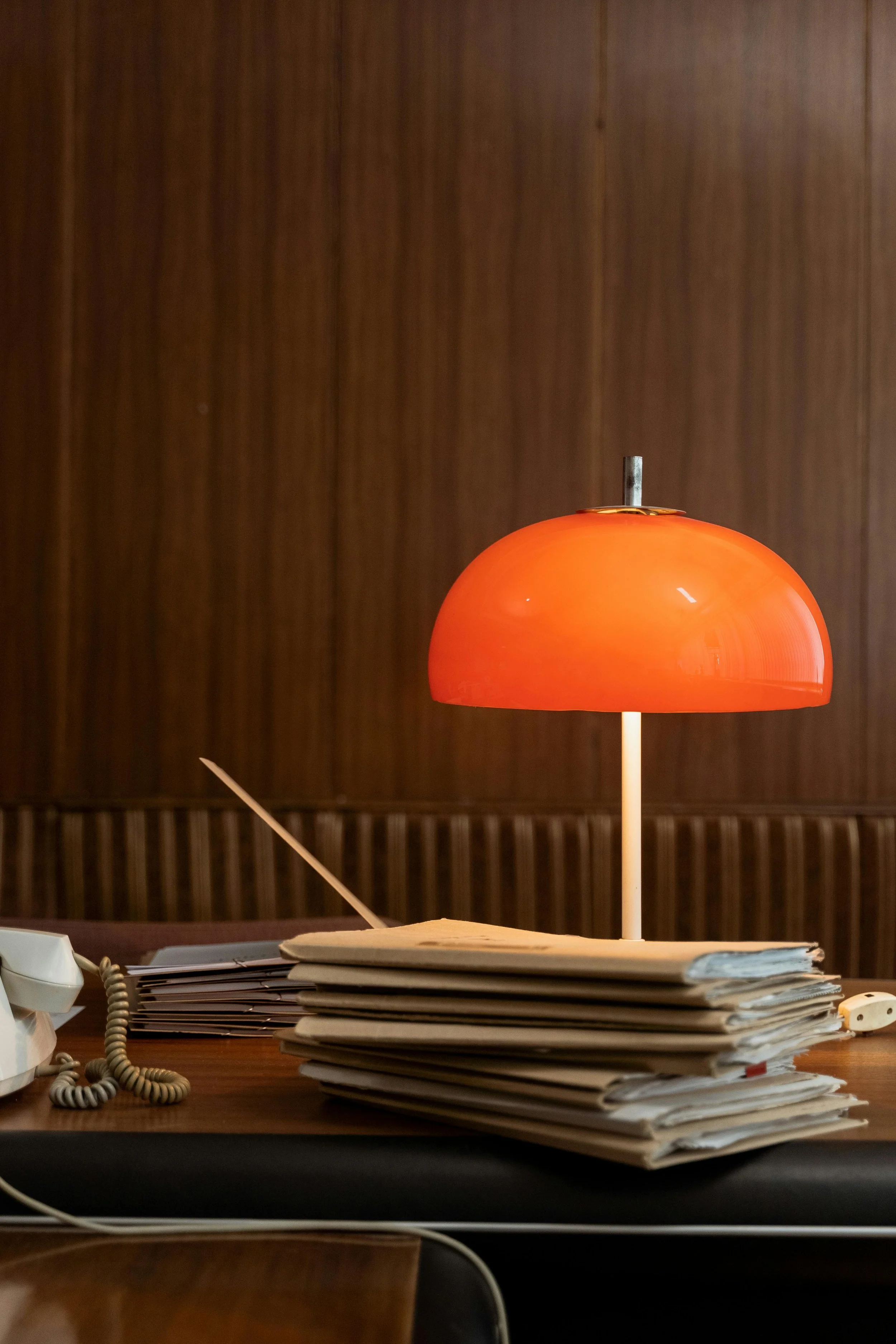 A cluttered desk with a stack of papers, a vintage beige rotary phone, an orange desk lamp, and a pen. The background features wooden paneling.