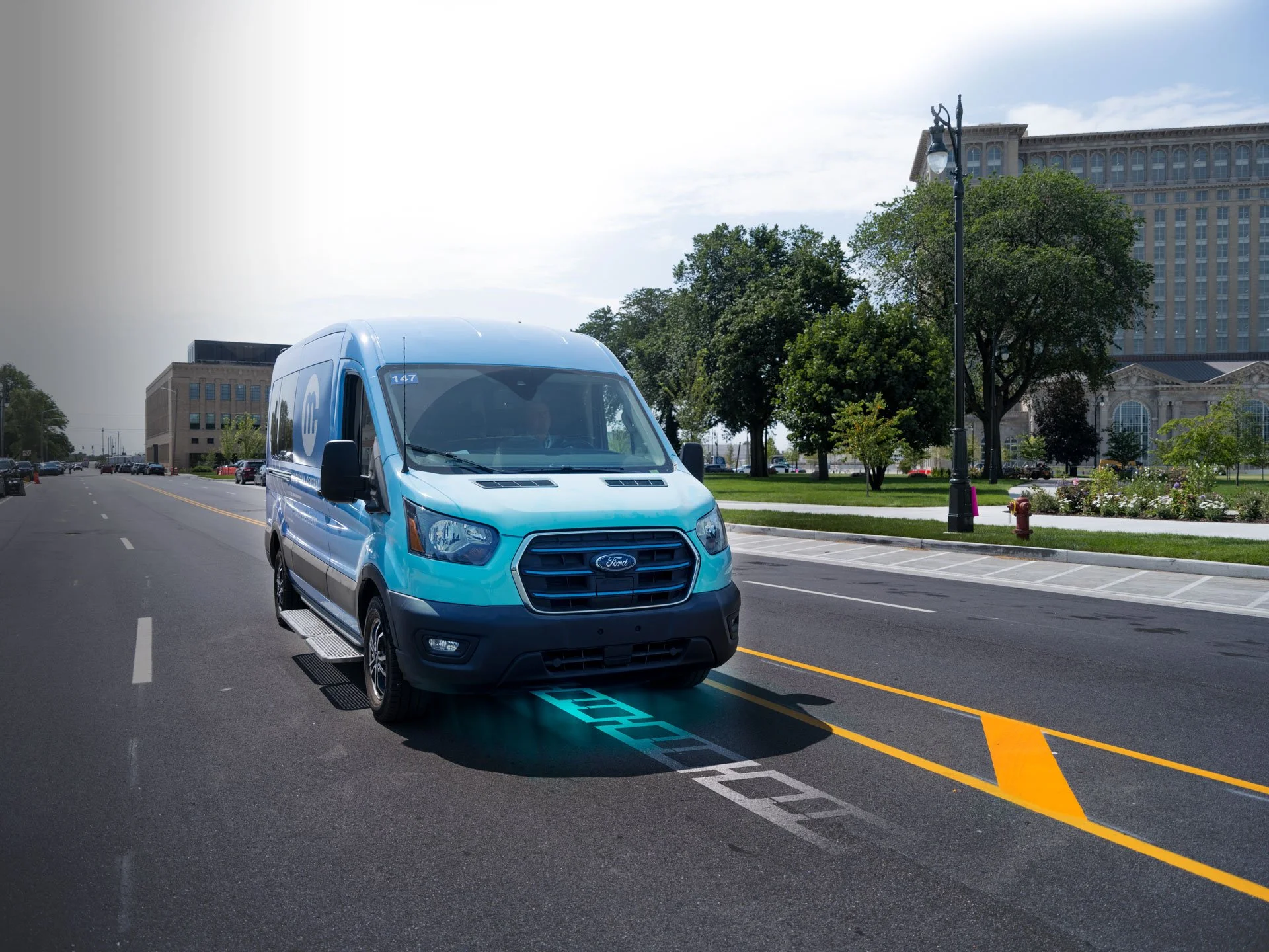 A blue commercial Ford van parked on a city street with a man at a charging station, surrounded by trees and a large building in the background.