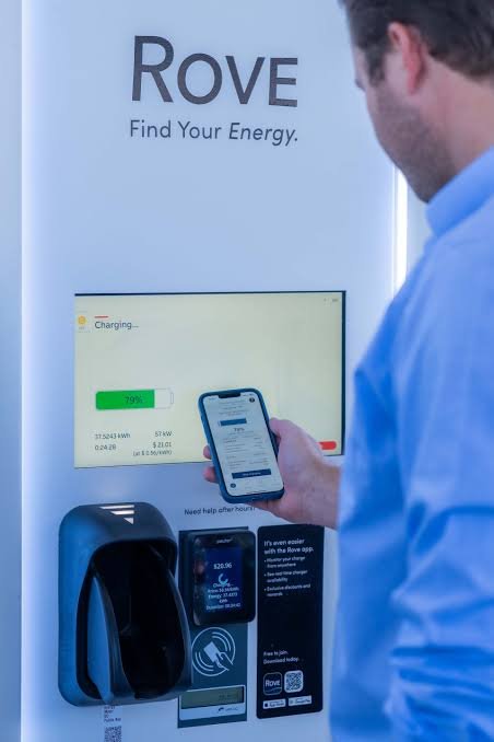 A man in a blue shirt using his smartphone to charge a device at a Rover electric vehicle charging station. The station displays a large screen with a green charging progress bar and the app interface. The Rover logo and tagline are visible on the wall.